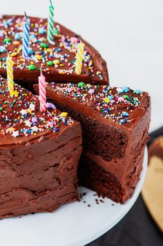 a chocolate cake with one slice cut out and lit candles on top, sitting on a white plate