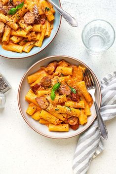 two bowls filled with pasta and meat on top of a white table next to silverware