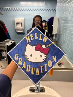 a person holding up a hello kitty graduation cap in front of a bathroom sink with blue tiles on the walls