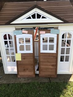 two wooden doors are open in front of a white shed with brown shingles on the roof