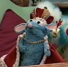 a stuffed mouse wearing a crown on top of a wooden table next to a red pillow