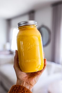 a person holding up a mason jar filled with yellow liquid in front of a bed