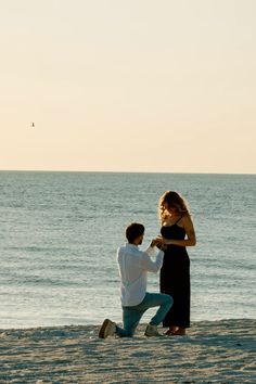 a man kneeling down next to a woman on top of a sandy beach near the ocean