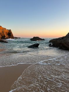 the beach is sandy and has waves coming in from the rocks on the shore as the sun sets