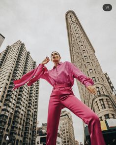 a woman in pink jumpsuits standing next to tall buildings with her arms outstretched