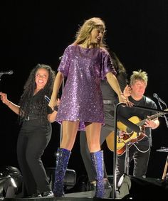 a woman in purple dress standing on stage with two men and one woman playing guitar