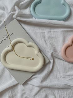 three ceramic clouds on a white sheet with a book and necklace in the foreground