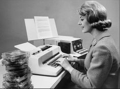 a woman sitting at a desk typing on an old typewriter with stacks of papers behind her