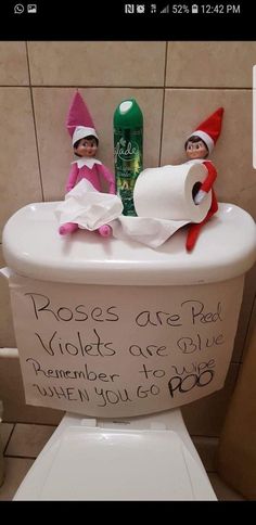 two elfs sitting on top of a toilet in a bathroom next to a roll of toilet paper