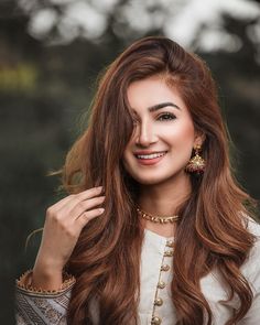 a woman with long brown hair wearing a white shirt and gold jewelry smiling at the camera