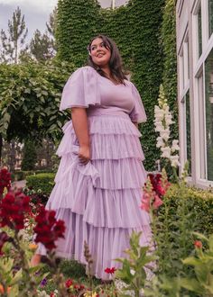 a woman wearing a purple dress standing in front of flowers and greenery with her hands on her hips