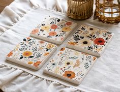 four square coasters with flowers on them sitting on a white tablecloth next to a wicker basket