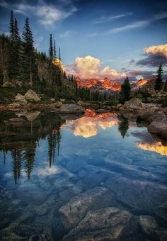 a lake surrounded by rocks and trees with the sun setting in the sky behind it