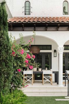 an outdoor dining area in front of a white house with pink flowers on the table