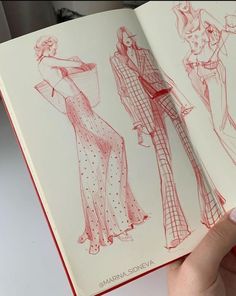 a hand holding an open book with sketches of women's clothing on the pages