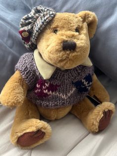 a teddy bear with a sweater and hat on it's head sitting on a bed