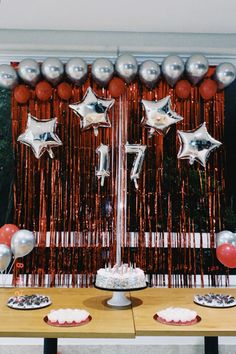 a table topped with cake and balloons next to a wall covered in red, silver and white streamers