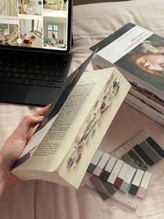 a person is holding an open book in front of a laptop and books on a bed