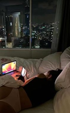 a person laying on a bed with a laptop in front of them and a view of the city at night