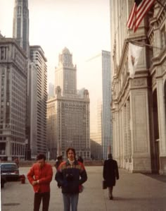 three people are standing on the sidewalk in front of tall buildings and an american flag