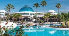 an outdoor swimming pool surrounded by palm trees and umbrellas in front of a domed glass building