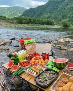 a picnic table with food and drinks on the side of a mountain river in front of some mountains