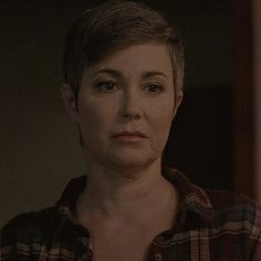 a woman with short hair wearing a plaid shirt looking at the camera while standing in front of a mirror