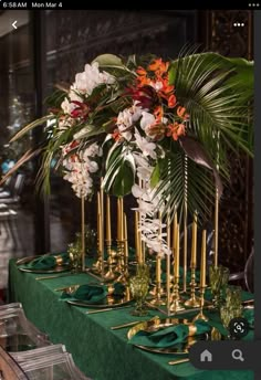 an arrangement of flowers and greenery on a table with gold candlesticks in the center