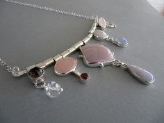 "Here is a stunning necklace made with sterling silver and nine stones. The stones are in fine and sterling silver settings and are soldered to sections of tubing that move freely on a heavy silver wire. From left to right: Smoky quartz in a tube setting with a faceted crystal. Rose quartz and 5mm tourmaline Square Phosposiderite in a serrated setting with little balls on each side. Teardrop shaped blue lace agate . Coin pearl with blue chalcedony Rose quartz oval with curl The center bar is 4\" Modern Silver Necklace With Stones, Sterling Silver Multi-stone Necklaces, Nine Stones, Tube Setting, Night At The Opera, Bar Jewelry, Montana Agate, Coin Pearls, Crystal Rose