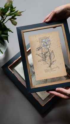 a person holding up a framed flower in front of a vase with flowers on it