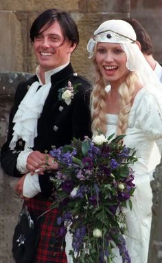 a man and woman dressed in scottish garb standing next to each other holding flowers