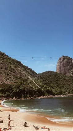 people are on the beach and in the water near some hills, with one person flying a kite