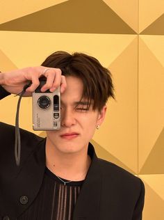 a man taking a selfie with his camera and wearing a black suit jacket, standing in front of a yellow geometric wall