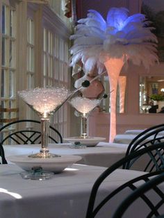 two martini glasses are sitting on a table in front of a tall vase with white feathers