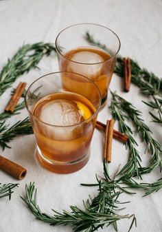 two glasses filled with drinks sitting on top of a table next to cinnamon sticks and rosemary