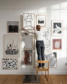 a woman standing on top of a wooden chair in front of a wall covered with art