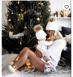 a woman sitting next to a christmas tree with her baby on her lap and she is wearing a white knitted hat