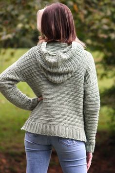 a woman standing in front of some trees wearing a sweater with a cowl neck