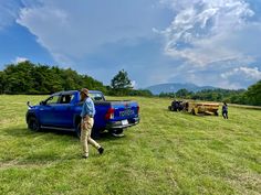 a man standing next to a blue truck on top of a lush green field with people in the background