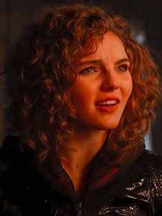 a close up of a person with curly hair wearing a black jacket and looking at the camera