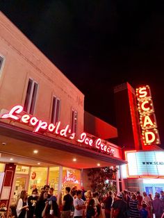 many people are standing outside of a theater at night with neon signs on the building