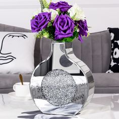 a silver vase filled with purple flowers on top of a white table next to a gray couch