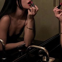 a woman sitting in front of a bathroom mirror while holding a lipstick brush and looking at her reflection