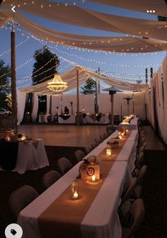 tables are set up with lit candles for an outdoor dinner party at night, surrounded by white draping and string lights