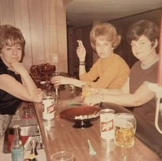 three women sitting at a table with drinks and cake in front of them, posing for the camera