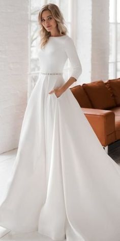 a woman standing in front of a couch wearing a white wedding dress with long sleeves