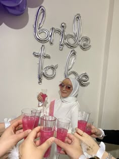 a group of people toasting with pink drinks in front of a bride to be balloon