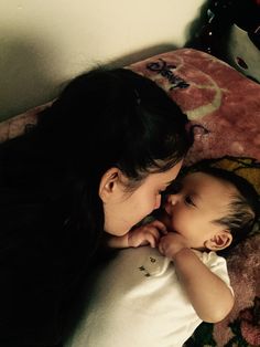 a woman laying on top of a bed next to a baby in her lap and touching her face