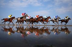 a group of jockeys riding horses across a wet beach next to the ocean on a sunny day