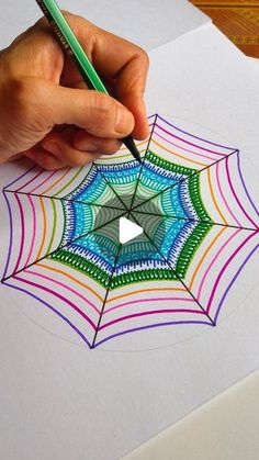 someone is drawing a spider web with colored pencils on top of the paper and holding a pen in their left hand